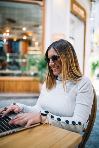 Mujer joven positiva que usa el ordenador portátil en la tabla en café de la calle