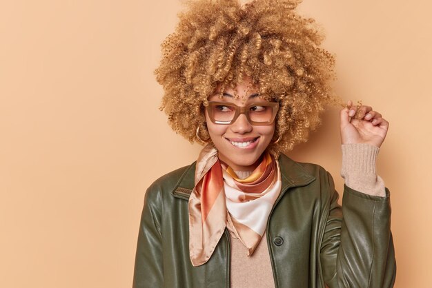 Mujer joven positiva riza el cabello, muerde los labios y mira hacia otro lado estando en buenos sueños sobre algo que usa gafas, una chaqueta de cuero de moda enfocada en poses contra el espacio de copia de fondo beige