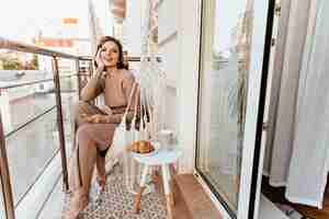 Foto gratuita mujer joven positiva en vestido largo sentado en el balcón con café y croissant. foto de niña rizada descalza disfrutando del desayuno en la terraza.