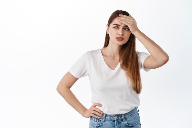 Mujer joven preocupada y angustiada que toca la frente mirando a un lado pensativa tiene un problema difícil situación de pie molesta y estresada contra el fondo blanco