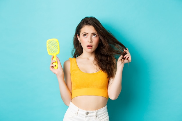 Mujer joven preocupada por la condición de su cabello, mostrando el pincel y mirando la esquina superior izquierda con rostro preocupado, de pie sobre fondo azul.