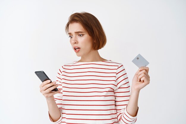 Mujer joven preocupada leyendo la pantalla del teléfono inteligente, sosteniendo una tarjeta de crédito de plástico, de pie ansiosa y confundida contra la pared blanca