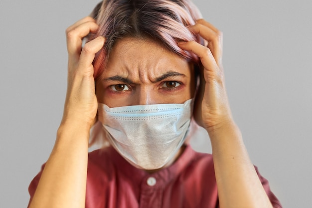Mujer joven preocupada en pánico que sufre de dolor de cabeza severo, con síntomas de COVID-19. Chica estresada en mascarilla médica preocupada por infecciones respiratorias contagiosas o gripe estacional