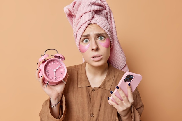 Mujer joven preocupada que se despierta tarde sostiene el despertador y el teléfono inteligente se somete a rutinas matutinas vestida con una toalla envuelta en pijama en la cabeza aislada sobre fondo marrón Concepto de despertar y descanso