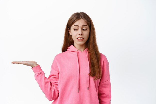 Mujer joven preocupada que se ve nerviosa por la palma vacía que sostiene el espacio de la copia del objeto de pie contra el fondo blanco