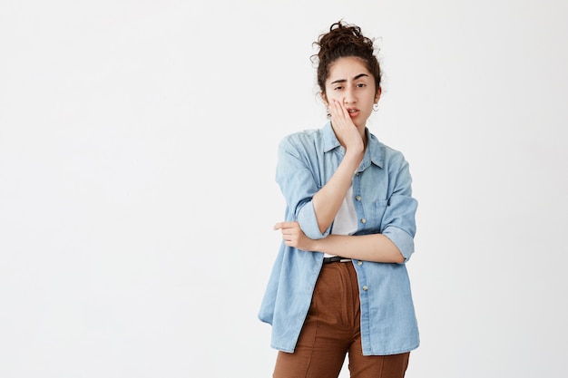 Foto gratuita mujer joven con problemas con cabello oscuro en moño en camisa de mezclilla tocando su mejilla y mirando hacia los lados con expresión dudosa y escéptica, tomando una importante decisión de vida, frunciendo el ceño