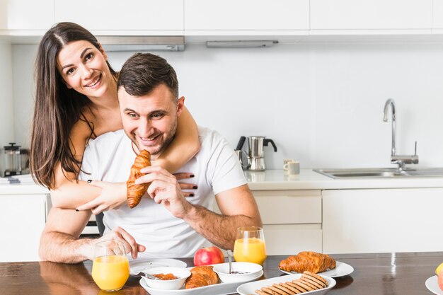 Mujer joven que abraza a su novio que desayuna en la cocina