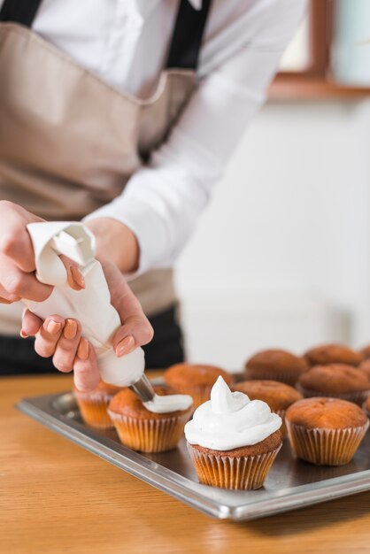 Mujer joven que adorna magdalenas con crema batida blanca apretando el bolso de la confitería