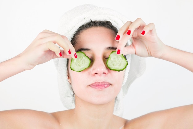 Mujer joven que aplica las rebanadas del pepino sobre sus ojos contra el fondo blanco