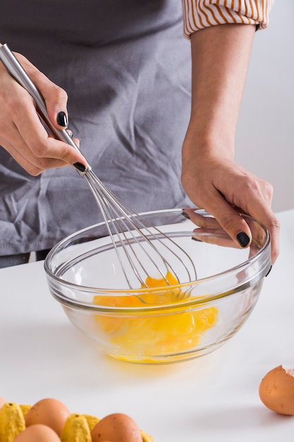 Mujer joven que bate la yema de huevo en el bol de vidrio con la batidora en la tabla blanca