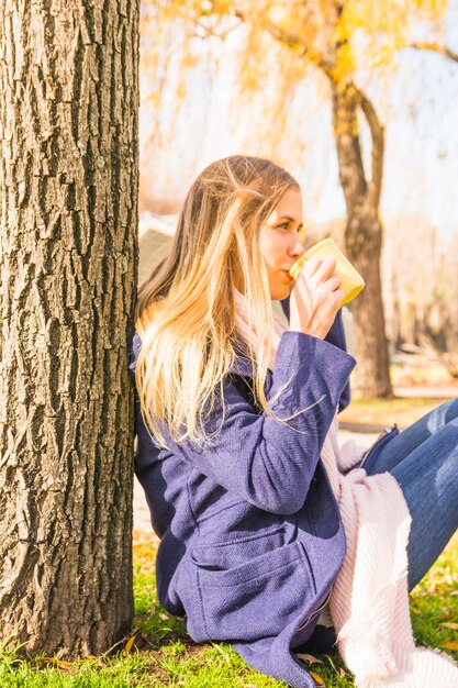 Mujer joven que bebe debajo de árbol en parque del otoño