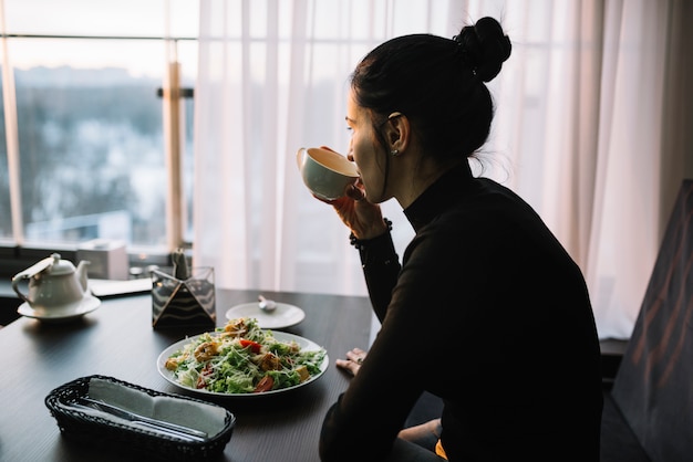 Foto gratuita mujer joven que bebe de la taza en la mesa con ensalada cerca de la ventana