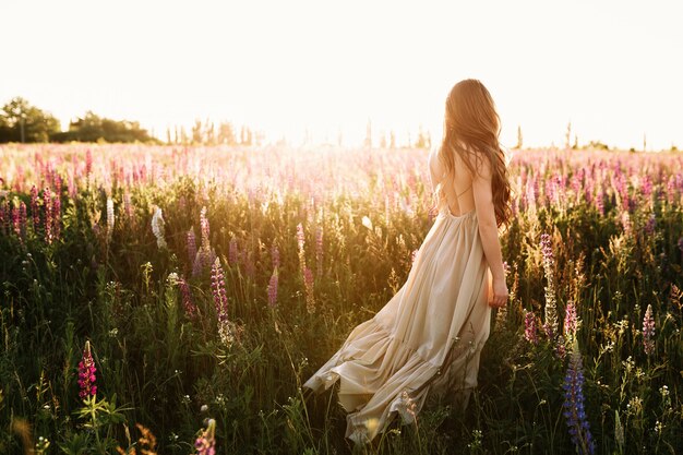 Mujer joven que camina en campo de flor en la puesta del sol en fondo.