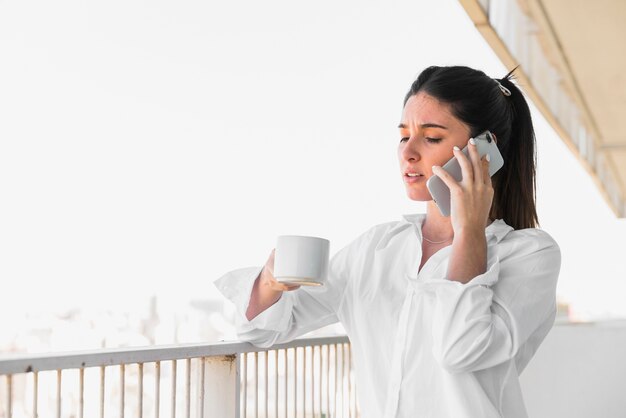 Mujer joven que se coloca en el balcón que sostiene la taza de café que habla en el teléfono móvil