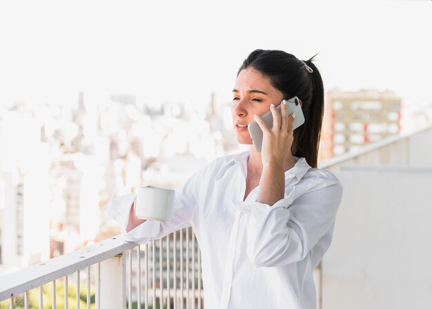 Mujer joven que se coloca en el balcón que sostiene la taza de café que habla en el teléfono móvil