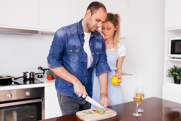 Mujer joven que se coloca cerca del marido que corta el bellpepper con el cuchillo en la tabla en la cocina