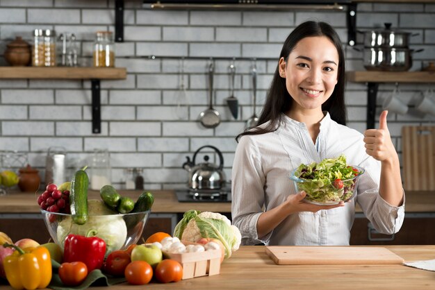 Mujer joven que se coloca cerca del mostrador de la cocina que muestra el pulgar encima de la muestra que sostiene la ensalada de las verduras