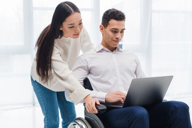 Mujer joven que se coloca detrás del hombre joven que se sienta en la silla de ruedas usando el ordenador portátil