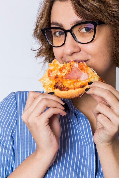 Mujer joven que come la pizza aislada en el fondo blanco que mira a la cámara