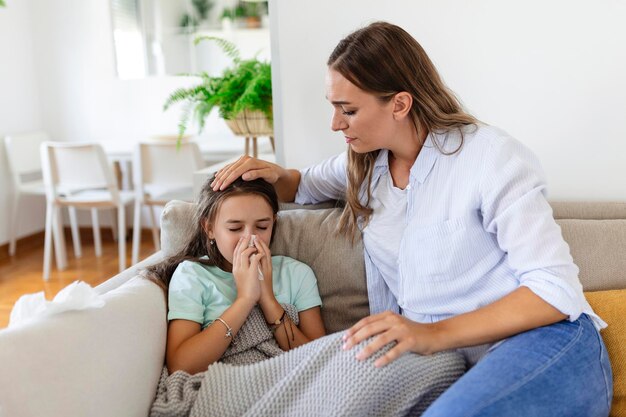 Mujer joven que controla la temperatura con la mano de la pequeña hija enferma Madre que controla la temperatura de su niña enferma Niño enfermo acostado en la cama debajo de la manta con la mujer que controla la fiebre en la frente con la mano