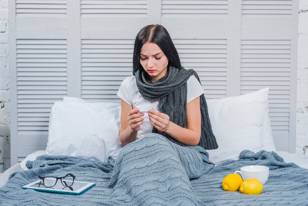 Mujer joven que controla temperatura en el termómetro que se sienta en cama con el limón; vaso; anteojos y tableta digital