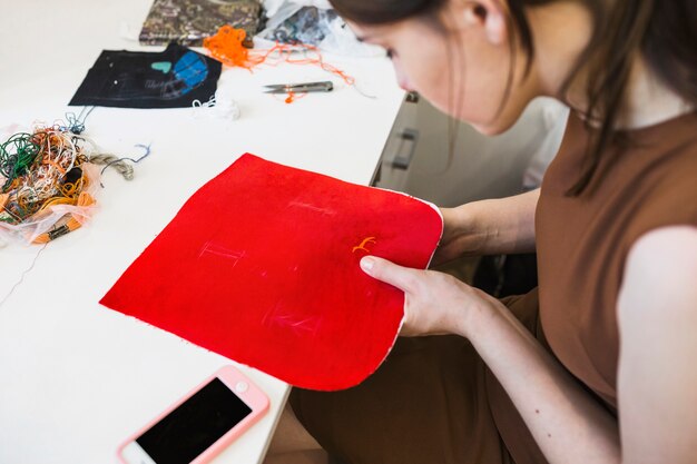 Mujer joven que cose el paño rojo con smartphone en el escritorio