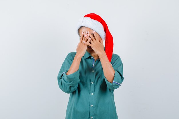 Mujer joven que cubre la cara con las manos en camisa, gorro de Papá Noel y parece deprimido. vista frontal.