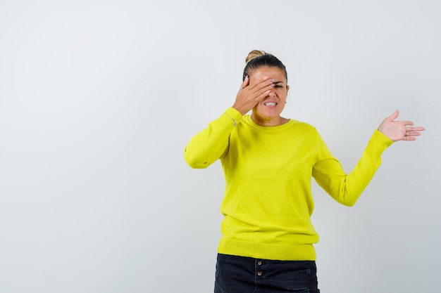 Mujer joven que cubre el ojo con la mano, estirando la mano hacia la derecha en suéter amarillo y pantalón negro y mirando feliz