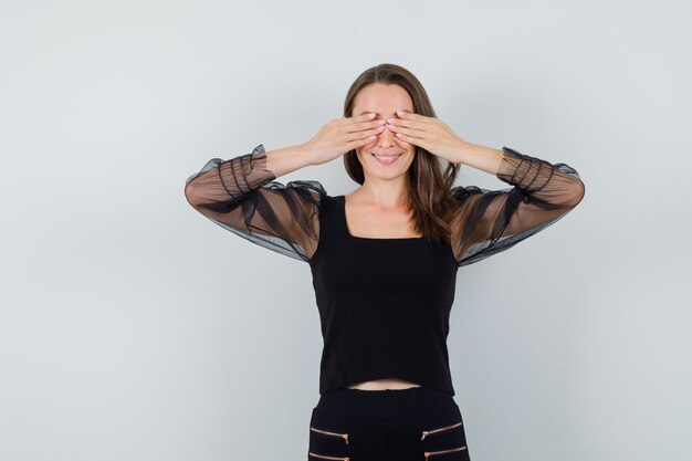 Mujer joven que cubre los ojos con ambas manos en blusa negra y pantalón negro y parece feliz. vista frontal.