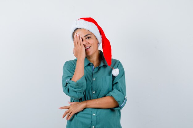 Mujer joven que cubre los ojos con la mano en la camisa, gorro de Papá Noel y mirando alegre. vista frontal.