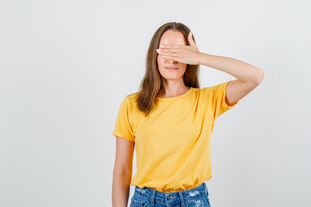 Mujer joven que cubre los ojos con la mano en camiseta, vista frontal de pantalones cortos.