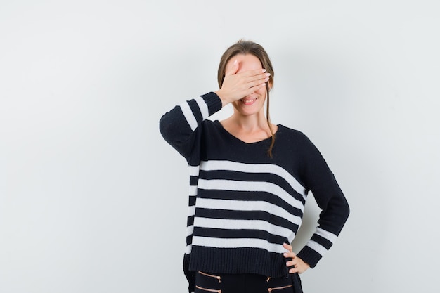 Mujer joven que cubre los ojos con las manos mientras sostiene las manos en la cintura en prendas de punto a rayas y pantalones negros y se ve feliz