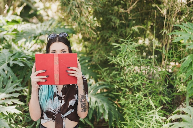 Foto gratuita mujer joven que cubre su boca con el libro que se coloca delante de las plantas crecientes