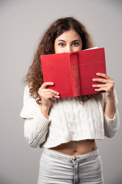 Mujer joven que cubre su rostro con un libro rojo sobre una pared gris. Foto de alta calidad