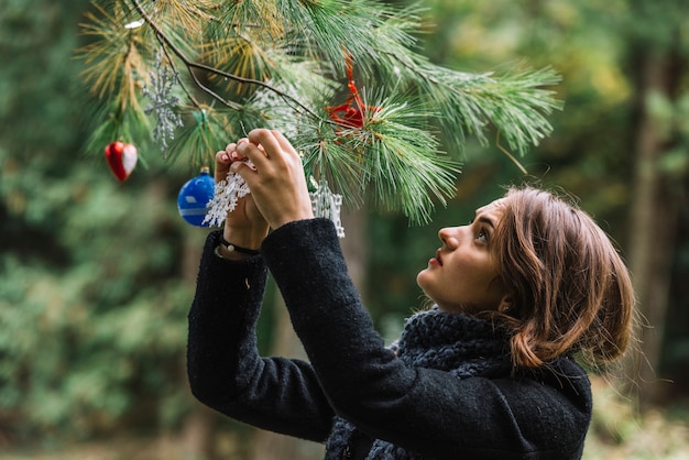 Mujer joven que cuelga los juguetes de la Navidad en la ramita en bosque