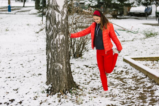 Mujer joven que estira su pierna debajo del árbol en invierno