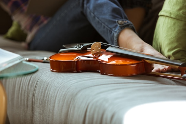 Mujer joven que estudia en casa durante cursos en línea o información gratuita por sí misma. Se convierte en músico, violinista mientras está aislado, cuarentena contra la propagación del coronavirus. Usando computadora portátil, teléfono inteligente.