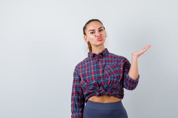 Mujer joven que finge mostrar algo mientras hace pucheros con los labios en camisa a cuadros, pantalones y se ve linda, vista frontal.