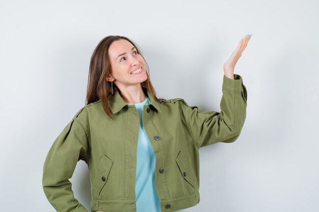 Mujer joven que finge mostrar algo mientras mira hacia arriba en chaqueta verde y se ve jovial. vista frontal.