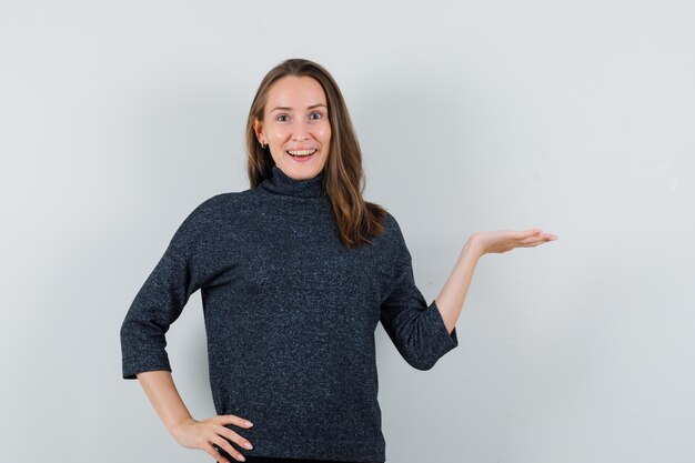 Mujer joven que finge sostener o mostrar algo en camisa y parece feliz. vista frontal.