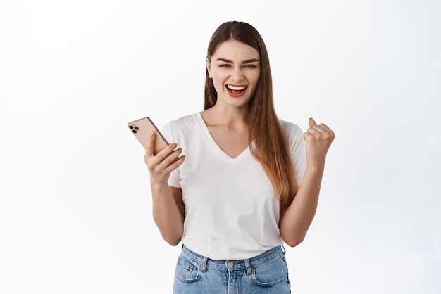 Mujer joven que gana en el teléfono dice sí regocijándose y sonriendo complacida sosteniendo el teléfono inteligente triunfando del logro de la aplicación en el teléfono celular de pie satisfecho contra el fondo blanco