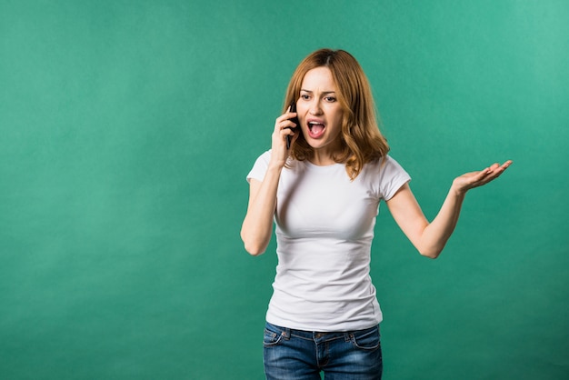 Mujer joven que grita en el teléfono elegante contra el contexto verde