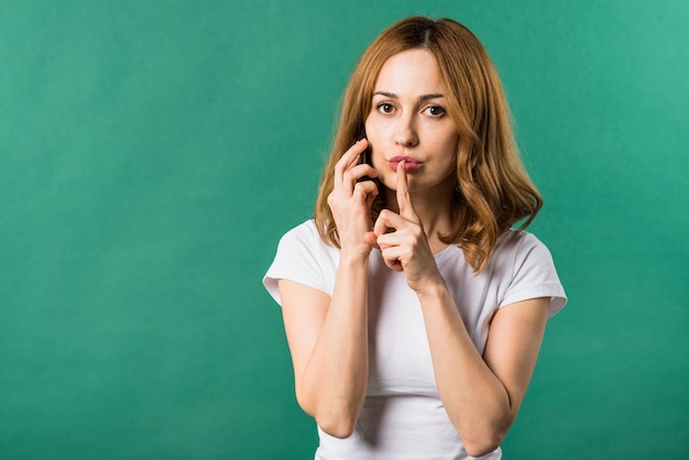 Foto gratuita mujer joven que habla en el teléfono móvil que hace gesto del silencio contra fondo verde