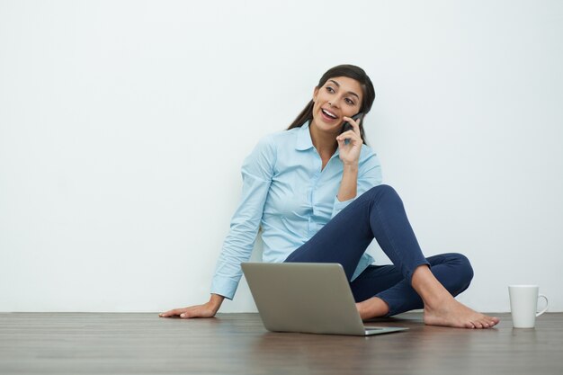 Mujer joven que habla en el teléfono en el piso con portátil