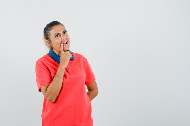 Mujer joven que se inclina el dedo cerca de la boca, de pie en pose de pensamiento en camiseta roja y mirando pensativo. vista frontal.