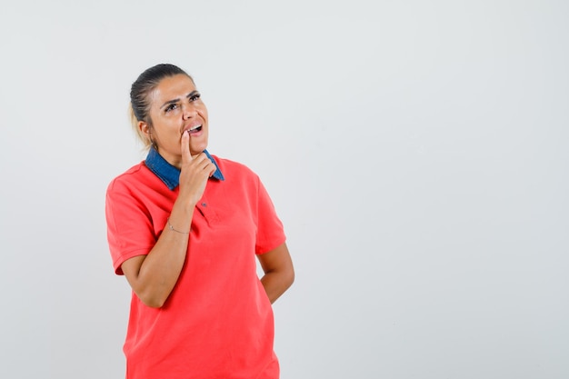 Mujer joven que se inclina el dedo cerca de la boca, de pie en pose de pensamiento en camiseta roja y mirando pensativo. vista frontal.