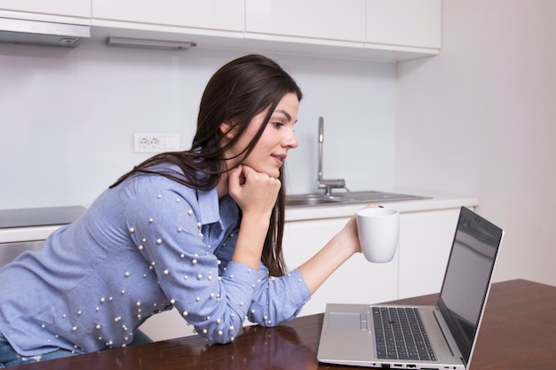 Mujer joven que se inclina en la tabla que mira la tableta digital que sostiene la taza de café disponible