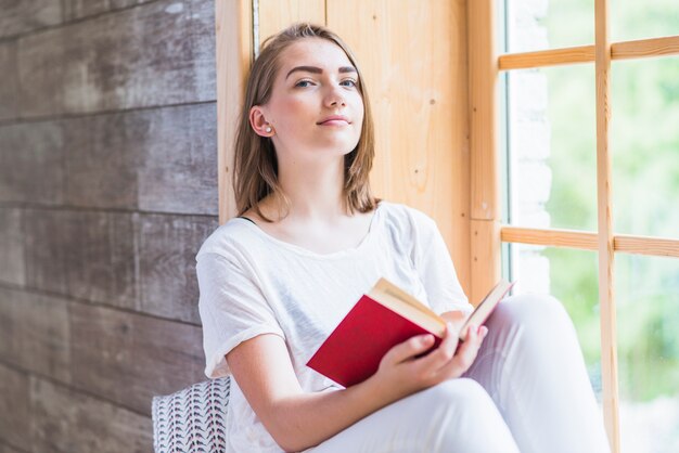 Mujer joven que se inclina en la ventana que sostiene el libro