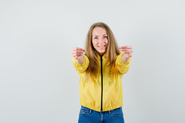 Mujer joven que invita a venir con chaqueta de bombardero amarilla y jean azul y luciendo optimista, vista frontal.