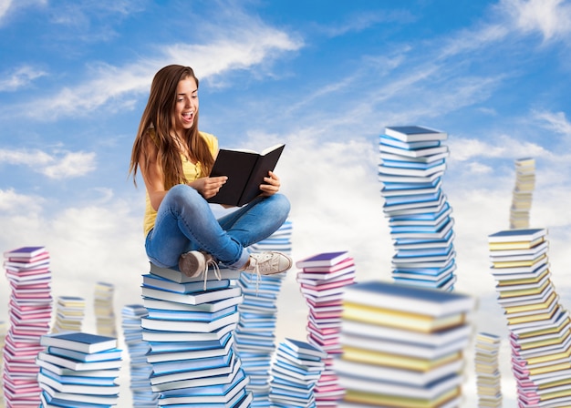 Mujer joven que lee un libro que se sienta en una pila de libros en el cielo